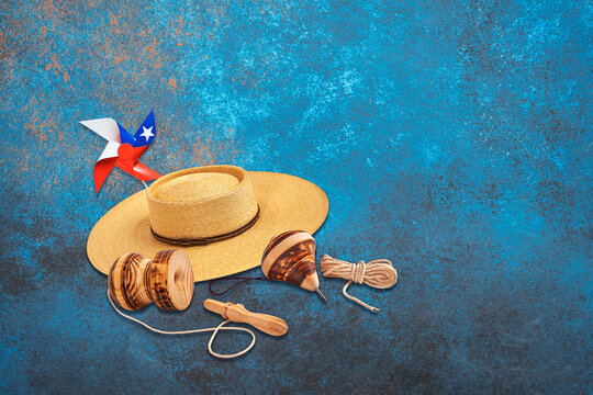 Chilean traditional toys, chupalla hat, and flag pinwheel on a blue textured background