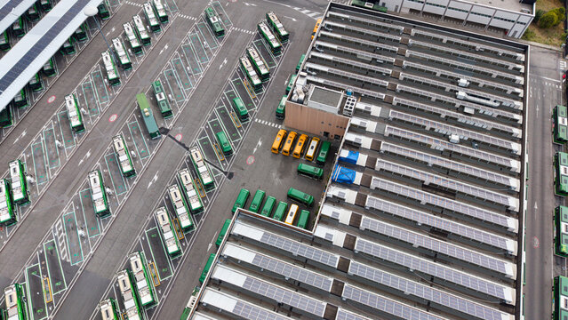 Aerial view of a modern public transport facility with multiple electric buses parked in rows and solar panels installed on the roof of the depot in San Donato Milanese, Lombardy, Italy.