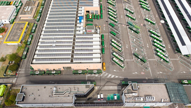 Aerial view of a modern public transport facility featuring rows of green electric buses parked at a depot with solar panels on the roof in San Donato Milanese, Lombardy, Italy.
