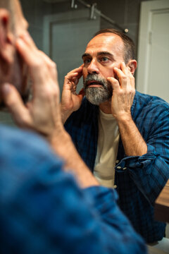 Bearded middle-aged man checking face in bathroom mirror