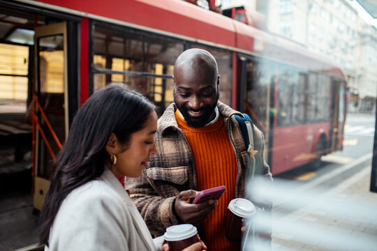 Two commuters with coffee using smartphone at city tram stop