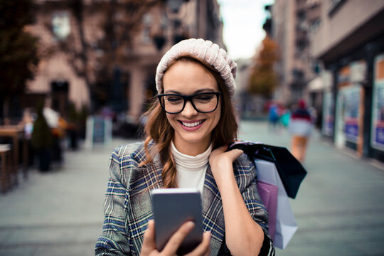 Smiling woman with shopping bags using smartphone on city street