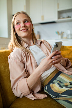 Smiling woman with under eye patches using smartphone at home on sofa