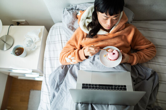 Woman eating dessert and using laptop in bed at home