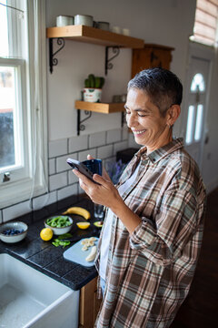 Smiling mature woman using smartphone in home kitchen preparing breakfast