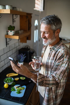 Mature man using smartphone and drinking water in home kitchen