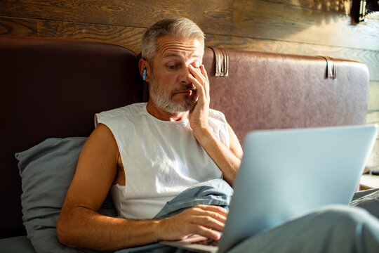 Tired middle-aged man working on laptop in bed at home