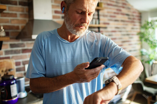 Active senior man checking smartwatch and phone in home kitchen
