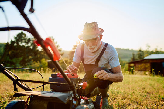 Senior man in overalls checking the engine oil level of a push lawn mower in a backyard at sunset