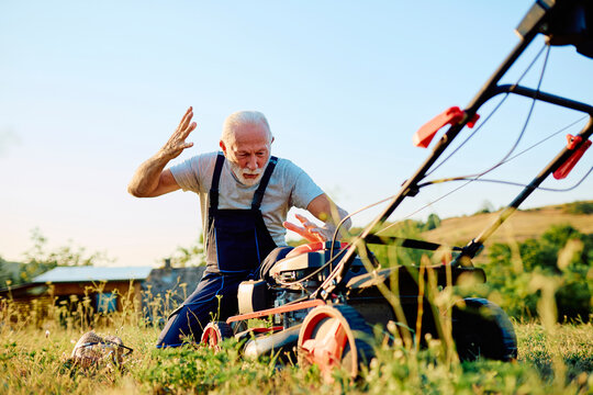 Senior gardener expressing frustration while kneeling on the grass, attempting to repair a broken lawnmower outdoors