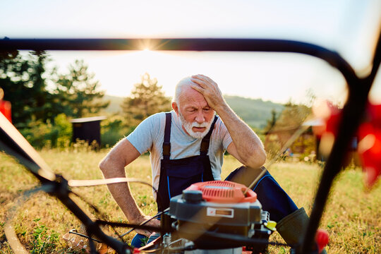 Older man experiencing stress and frustration, holding head while trying to repair a broken lawn mower in an outdoor garden at sunset