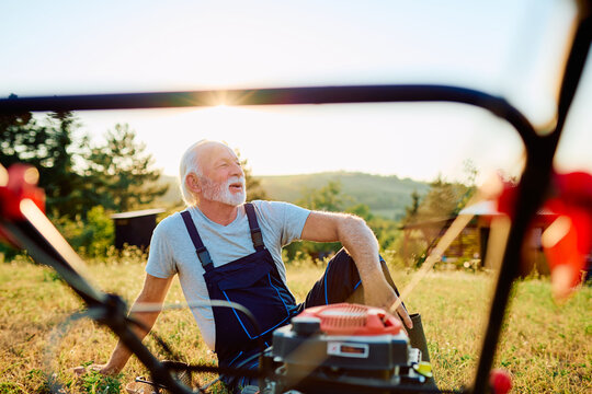 Senior man in overalls resting next to a garden tiller machine in a field, enjoying the sunset after working