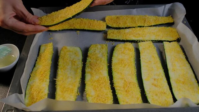 Hands coat zucchini slices with breadcrumbs on parchment-lined baking sheet in Cooking kitchen, overhead view.