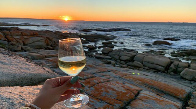 Hand holds glass of white wine at sunset overlooking a rocky coastline.