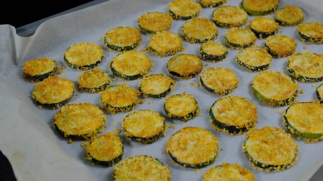 Sliced zucchini rounds coated in yellow breadcrumbs arranged on parchment paper over a baking pan, ready for oven cooking in a kitchen preparation area during daytime.