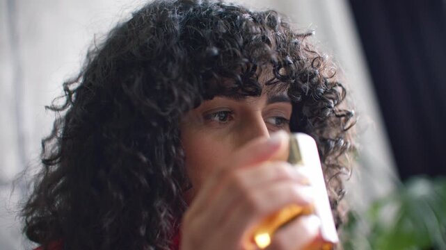 Close up of young Caucasian woman smiling while looking at laptop screen near window. Female with curly dark hair resting chin on hand. Enjoying online conversation or remote work indoors.