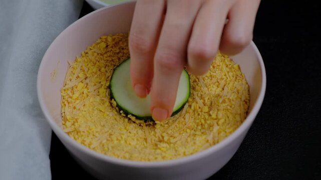 Close-up overhead of hands coating cucumber slices in breadcrumbs inside a pink bowl, set against a black background, during food preparation in Cooking.