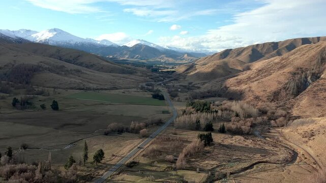 Aerial view of Burkes Pass with a road through a valley surrounded by snow-capped mountains and rolling hills in Burkes Pass, Canterbury Region, New Zealand.