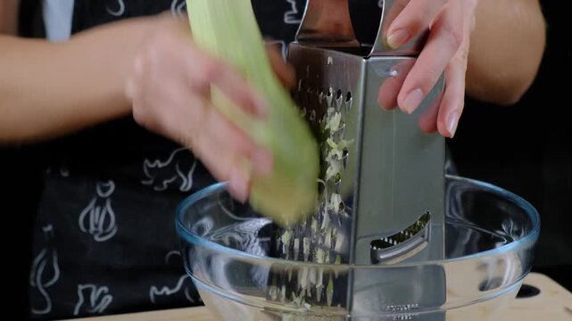 Adult hands grating fresh celery with metal grater into glass bowl on wooden surface in Cooking kitchen, daytime.