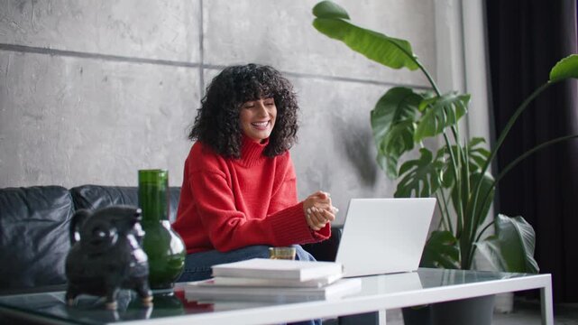 Young Caucasian woman having video call on laptop while sitting in modern living room. Female with curly dark hair gesturing during online meeting. Remote work communication from cozy home office.