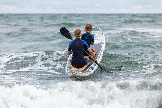 Two siblings boy girl paddle SUP board stormy Baltic waves, wearing wetsuits and sharing steady effort. Energetic seaside scene conveys fun, teamwork, caution and summer adventure on Ostsee coast