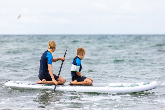 Two siblings boy girl paddle SUP board stormy Baltic waves, wearing wetsuits and sharing steady effort. Energetic seaside scene conveys fun, teamwork, caution and summer adventure on Ostsee coast