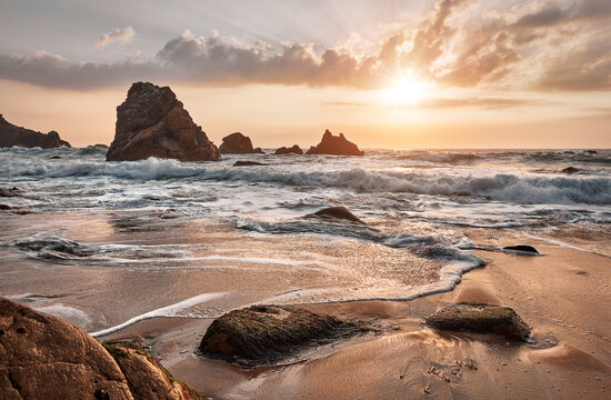 Sand beach among rocks on evening sunset. Ursa Beach near Cape Roca (Cabo da Roca) at Atlantic Ocean coast in Portugal. Summer landscape. Beach with splashes of waves crashing of stone rocks.