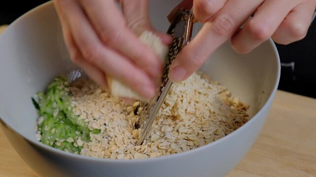 Adult hands mix rolled oats, chopped greens, and grated cheese in a white bowl on a wooden counter during cooking.
