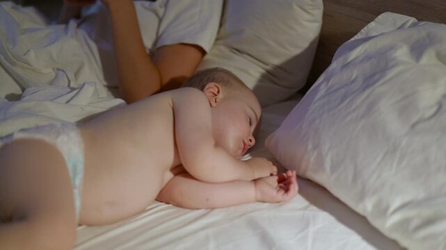 Baby lying on bed with white sheets and pillows, peacefully sleeping beside an adult, showcasing a serene nighttime environment in a cozy bedroom setting