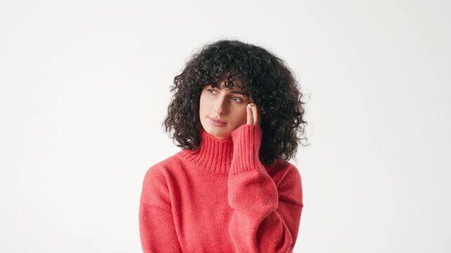Thoughtful young Caucasian woman touching temple while looking away. Female with curly dark hair wearing red sweater. Pensive portrait expressing reflection decision making and concentration.