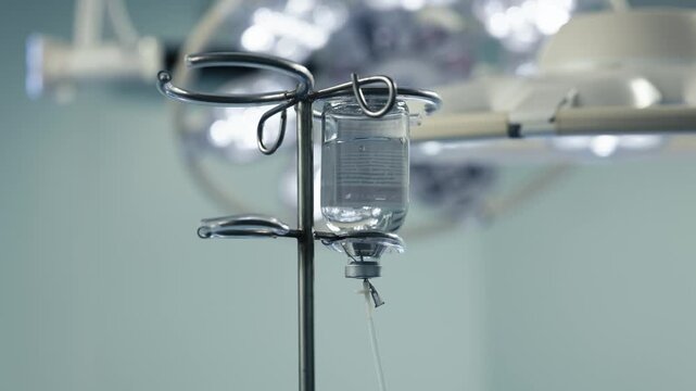 Bottle with clear infusion solution attached to an IV pole in an operating room. The system is prepared for intravenous fluid administration to a patient.