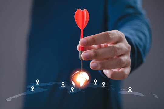 Business hand placing a red dart on a glowing world map target, symbolizing global strategy, market targeting, business growth, international expansion, location planning, success, goal achievement.