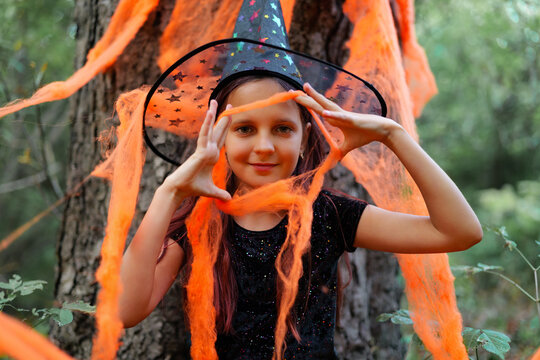 A girl wearing a witch hat stands near a tree, holding strands of orange fabric. It is Halloween time, and the area is decorated for the holiday