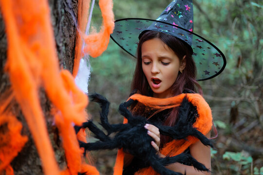 A young girl in a witch hat stands in a wooded area wrapped in orange decorations. She is surprised by a big toy spider in her hands. Trees surround her