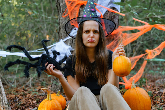 A person sits on the ground in a forest, wearing a witch hat. They hold a black spider toy and two pumpkins. Halloween decorations are in the background