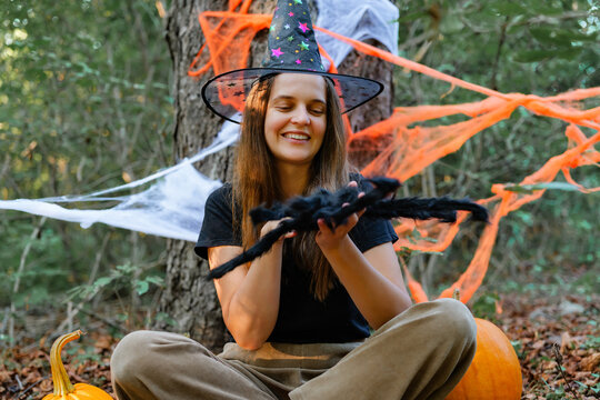 A girl sits on the ground in a forest wearing a witch hat. She holds a fake spider with pumpkins and Halloween decorations around her