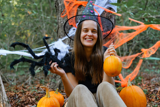 A woman wearing a witch hat is sitting in a forest. She holds a small pumpkin in one hand and a toy spider in the other. There are orange decorations around her