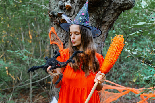 A girl in a red dress and witch hat stands in a forest. She holds a broom in one hand and a toy spider in the other. Orange decorations hang from trees
