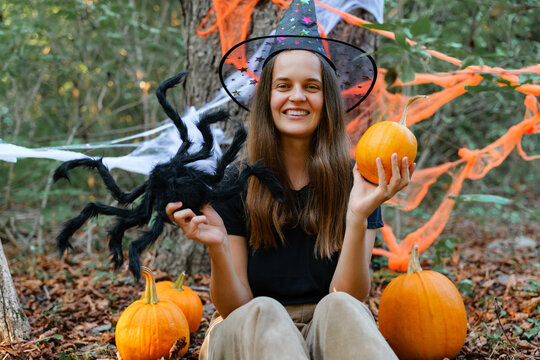 A woman sits in a forest surrounded by pumpkins and Halloween decorations. She holds a pumpkin in one hand and a toy spider in the other
