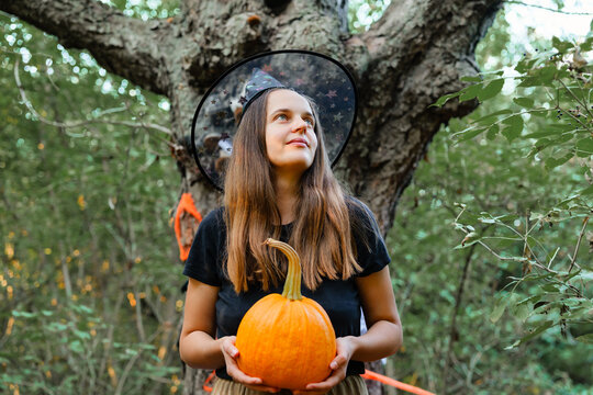 A woman stands in a forest with trees around her. She holds a pumpkin and looks up with a smile. The sun shows through the leaves