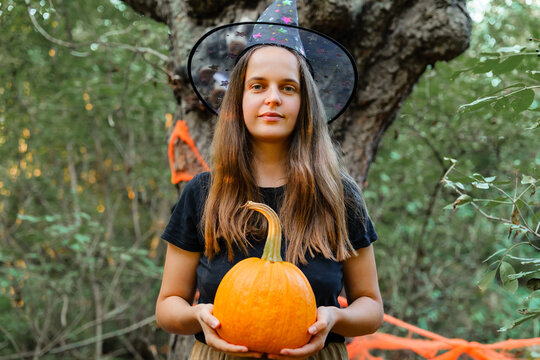 A woman stands in a forest wearing a witch hat and holding a pumpkin. The setting includes Halloween decorations and trees around her