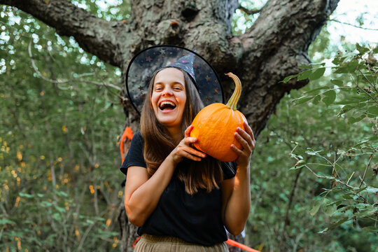 A woman stands in a forest, smiling as she holds a bright orange pumpkin. She wears a witch hat and enjoys the fall season among the trees