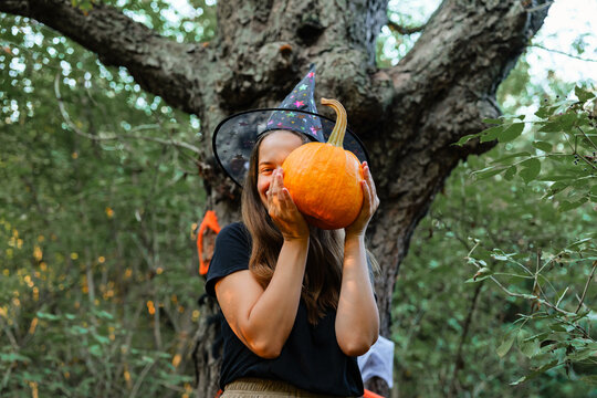 A woman is smiling and holding a bright orange pumpkin with both hands. She stands in a wooded area surrounded by trees