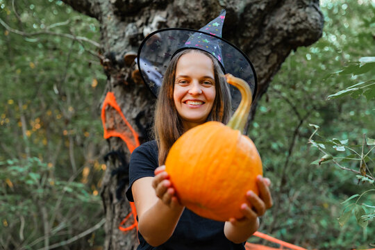 A girl stands in a forest showing a bright orange pumpkin. She wears a witch hat and smiles. Trees surround her, creating an autumn scene
