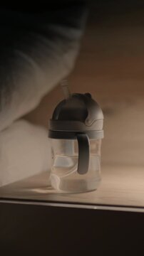 Transparent baby sippy cup with gray lid and straw placed on wooden surface, soft lighting highlighting the cup's features and surrounding elements