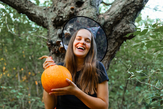 In a forest during autumn, a person smiles while holding a pumpkin and wearing a witch hat. Trees surround the scene, creating a festive atmosphere