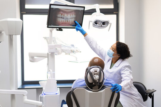 Black female dentist explaining digital dental images to male patient on monitor before treatment, demonstrating modern technology, patient education and personalized oral care