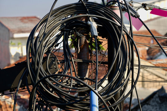 Coiled Black Cables on Utility Pole A bundle of black fiber optic and communication cables tightly coiled around a utility pole outdoors, overlooking urban residential rooftops with terracotta tiles.