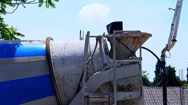 A cement mixer truck with a rusty rotating drum and metal chute sits parked outdoors near a tiled roof building, illustrating heavy equipment used in construction work.