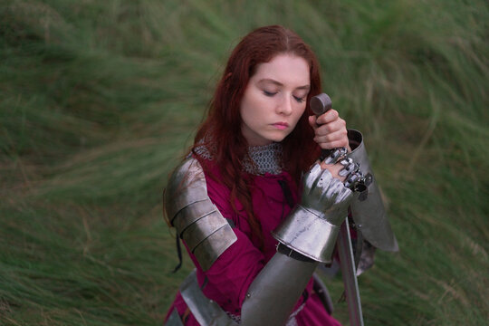 A young woman dressed as a medieval knight sits dejectedly on a grassy hillside, clutching her sword and helmet.